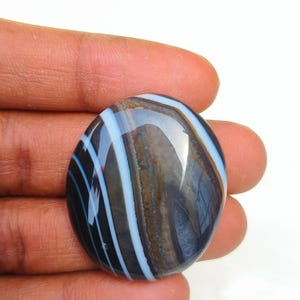Puede incluir: Una gema ovalada pulida con un llamativo patrón de bandas azules, blancas y marrones. La piedra tiene un acabado brillante y se sostiene en una mano, mostrando su belleza natural. Una pieza de joyería única.