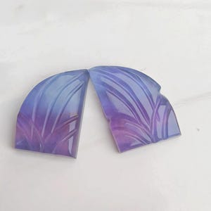 Könnte beinhalten: Zwei geschnitzte Edelsteinkabochons in Violett- und Blautönen. Die Cabochons sind halbkreisförmig und haben ein geschnitztes Blattdesign.