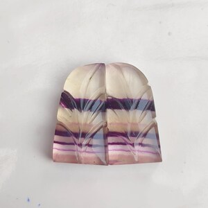 Könnte beinhalten: Ein Paar geschnitzte Fluorit-Cabochons mit einem geschichteten, gestreiften Muster in Violett-, Rosa- und Weißtönen. Die Cabochons sind mit einem Blattdesign geschnitzt.
