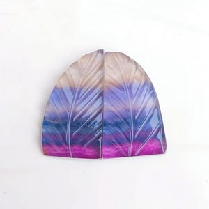 Könnte beinhalten: Zwei geschnitzte Edelsteinkabochons mit einem geschichteten, gefiederten Design. Die Cabochons sind eine Mischung aus Lila, Blau, Rosa und Beige.