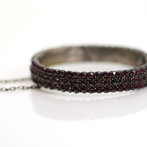 Könnte beinhalten: Ein silberfarbenes Armband mit Kettchen, besetzt mit Reihen kleiner, dunkelroter Edelsteine. Das Schmuckstück liegt vor weißem Hintergrund, wodurch die Details und die Farbe hervorgehoben werden.