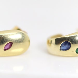 Puede incluir: Un par de pendientes de aro de oro, cada uno con tres piedras preciosas. Las piedras preciosas son una esmeralda verde, un zafiro azul y un rubí rojo.