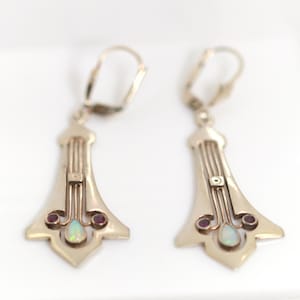Könnte beinhalten: Ein Paar Ohrringe im Art-Deco-Stil mit silberfarbenem Finish. Jeder Ohrring hat einen zentralen Stein, der einem Opal ähnelt, flankiert von zwei kleinen roten Steinen. Das Design umfasst vertikale Linien und einen Klappbügelverschluss.