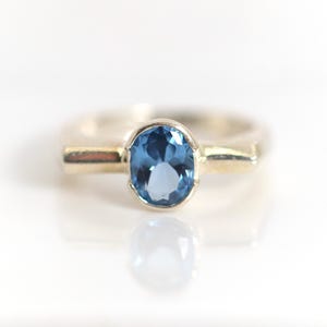 Puede incluir: Un anillo de plata con una gema azul grande de forma ovalada. La gema está engastada en un bisel y la banda del anillo tiene un diseño simple y liso. El anillo se muestra sobre una superficie blanca, con un reflejo debajo.