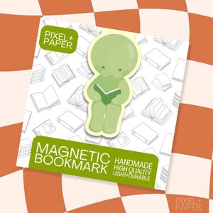 May include: A light green magnetic bookmark featuring a cartoon character reading a book. The bookmark is displayed on a white card with a repeating line-art pattern of books and the text PIXEL + PAPER and MAGNETIC BOOKMARK HANDMADE.