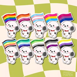 Puede incluir: Un conjunto de diez pegatinas troqueladas que muestran un perro de dibujos animados blanco con un collar rojo y manchas negras, cada uno sosteniendo una bandera de orgullo diferente. Las banderas incluyen diseños arcoíris, lésbico, transgénero, pansexual, bisexual, poliamoroso, gay, arromántico, asexual y no binario.