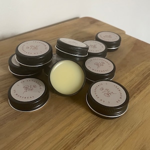 Puede incluir: Primer plano de varios botes pequeños, redondos y negros con etiquetas blancas. Las etiquetas presentan un diseño floral y el texto "La Savonnerie de Montreal". Un bote está abierto, revelando un bálsamo amarillo pálido en su interior.