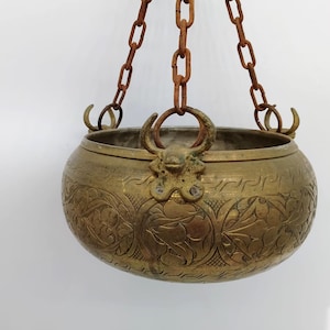 May include: A brass hanging planter with intricate floral designs and a rustic chain. The planter has a rounded shape and a wide opening.