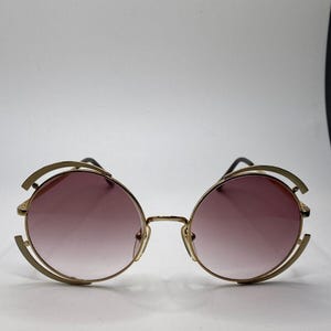 May include: Round sunglasses with a gold metal frame and gradient pink lenses. The sunglasses feature a distinctive design with curved accents on the top and sides of the frames. The lenses are a soft pink colour.
