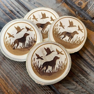 May include: Four wooden coasters with a laser-etched design of a dog and birds. The coasters are round and have a natural wood finish.