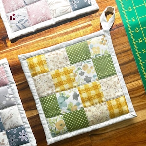 May include: Two quilted pot holders with a patchwork design. One pot holder is yellow, green, and white with a floral pattern. The other pot holder is pink, grey, and white with a floral pattern.