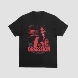 Puede incluir: Una camiseta negra de manga corta con un estampado gráfico rojo de una persona sonriendo, una casa y un coche. El texto dice: BE CAREFUL WHO YOU WISH FOR... OBSESSION JUST BECAUSE YOU CHOSE THIS FOR HER DOESN'T MAKE IT LESS REAL A FILM BY CURRY BARKER.