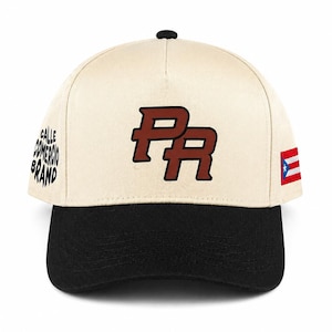 May include: A cream-coloured baseball cap with a black brim and a small black button on top. The front features the letters PR in dark red with black outlines. The right side displays a small Puerto Rican flag, and the left side has the text CALLE COMERCIO BRAND in black.