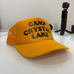 Puede incluir: Gorra de camionero amarilla con el texto "CAMP CRYSTAL LAKE" en negro. La gorra tiene una parte trasera de malla y una visera curva. La gorra está sobre una superficie blanca.