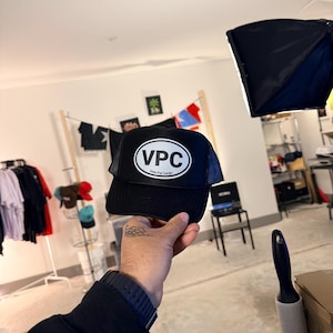 Puede incluir: Gorra de camionero negra con parte trasera de malla y visera curva. La parte delantera de la gorra presenta un parche ovalado blanco con las letras "VPC" en negrita, en negro. Debajo de las letras está el texto "Vote Pal Carajo".
