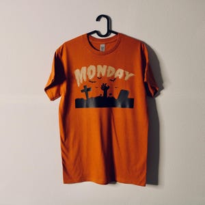Könnte beinhalten: Ein orangefarbenes T-Shirt mit einer schwarzen Grafik eines Friedhofs und dem Wort "MONDAY" in weißer Schrift.