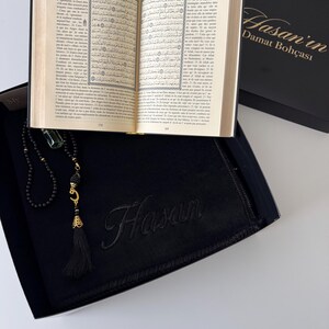 K&ouml;nnte beinhalten: Eine schwarze Geschenkbox mit einem aufgeschlagenen Buch mit arabischem Text, einer schwarzen Gebetskette mit goldfarbenem Halbmond-Anh&auml;nger und Quaste sowie einem schwarzen Stoffartikel, der mit dem Namen Hasan bestickt ist. Der Deckel tr&auml;gt den Text Hasan'in Damat Boh&ccedil;ası.