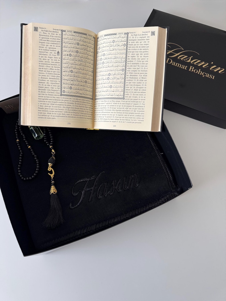 K&ouml;nnte beinhalten: Eine schwarze Geschenkbox mit einem aufgeschlagenen Buch mit arabischem Text, einer schwarzen Gebetskette mit goldfarbenem Halbmond-Anh&auml;nger und Quaste sowie einem schwarzen Stoffartikel, der mit dem Namen Hasan bestickt ist. Der Deckel tr&auml;gt den Text Hasan'in Damat Boh&ccedil;ası.