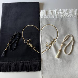 Puede incluir: Una alfombra de oración negra con flecos negros y una alfombra de oración blanca con flecos blancos. Cada alfombra presenta un contorno de corazón bordado en oro y un nombre, Sener en la negra y Sena en la blanca. Un rosario negro y uno de perlas blancas se colocan sobre las alfombras.
