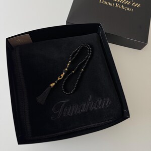 K&ouml;nnte beinhalten: Eine schwarze Geschenkbox mit einem schwarzen Samtstoff, der mit dem Namen Tunahan bestickt ist, sowie einer schwarzen Gebetskette mit goldfarbenen Akzenten und einer schwarzen Quaste. Der Deckel zeigt den Text Tunahan'in Damat Boh&ccedil;ası.
