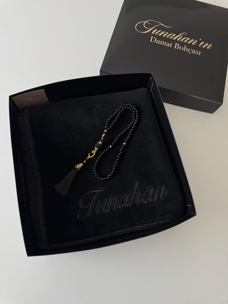 K&ouml;nnte beinhalten: Eine schwarze Geschenkbox mit einem schwarzen Samtstoff, der mit dem Namen Tunahan bestickt ist, sowie einer schwarzen Gebetskette mit goldfarbenen Akzenten und einer schwarzen Quaste. Der Deckel zeigt den Text Tunahan'in Damat Boh&ccedil;ası.