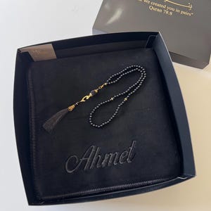 K&ouml;nnte beinhalten: Eine schwarze Gebetsmatte mit dem eingestickten Namen Ahmet, begleitet von schwarzen Gebetsperlen mit goldfarbenen Akzenten und einer Quaste. Das Set befindet sich in einer schwarzen Geschenkbox mit dem Text And We created you in pairs Quran 78:8.