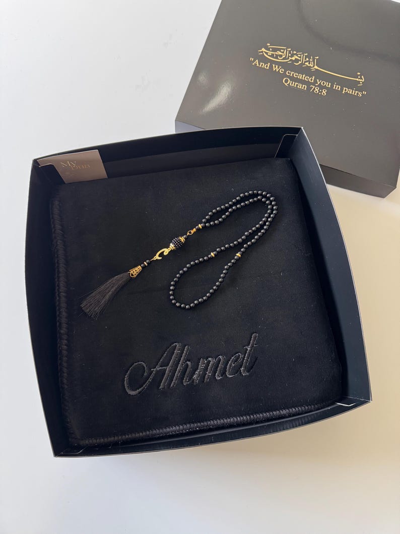 K&ouml;nnte beinhalten: Eine schwarze Gebetsmatte mit dem eingestickten Namen Ahmet, begleitet von schwarzen Gebetsperlen mit goldfarbenen Akzenten und einer Quaste. Das Set befindet sich in einer schwarzen Geschenkbox mit dem Text And We created you in pairs Quran 78:8.