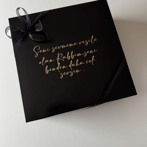 K&ouml;nnte beinhalten: Eine quadratische, schwarze Geschenkbox mit gl&auml;nzender Oberfl&auml;che, verziert mit einer schwarzen Schleife in der oberen linken Ecke. Der Deckel tr&auml;gt einen goldenen kursiven Schriftzug: Seni sevmeme vesile olan Rabbim, seni benden daha &ccedil;ok sevsin.