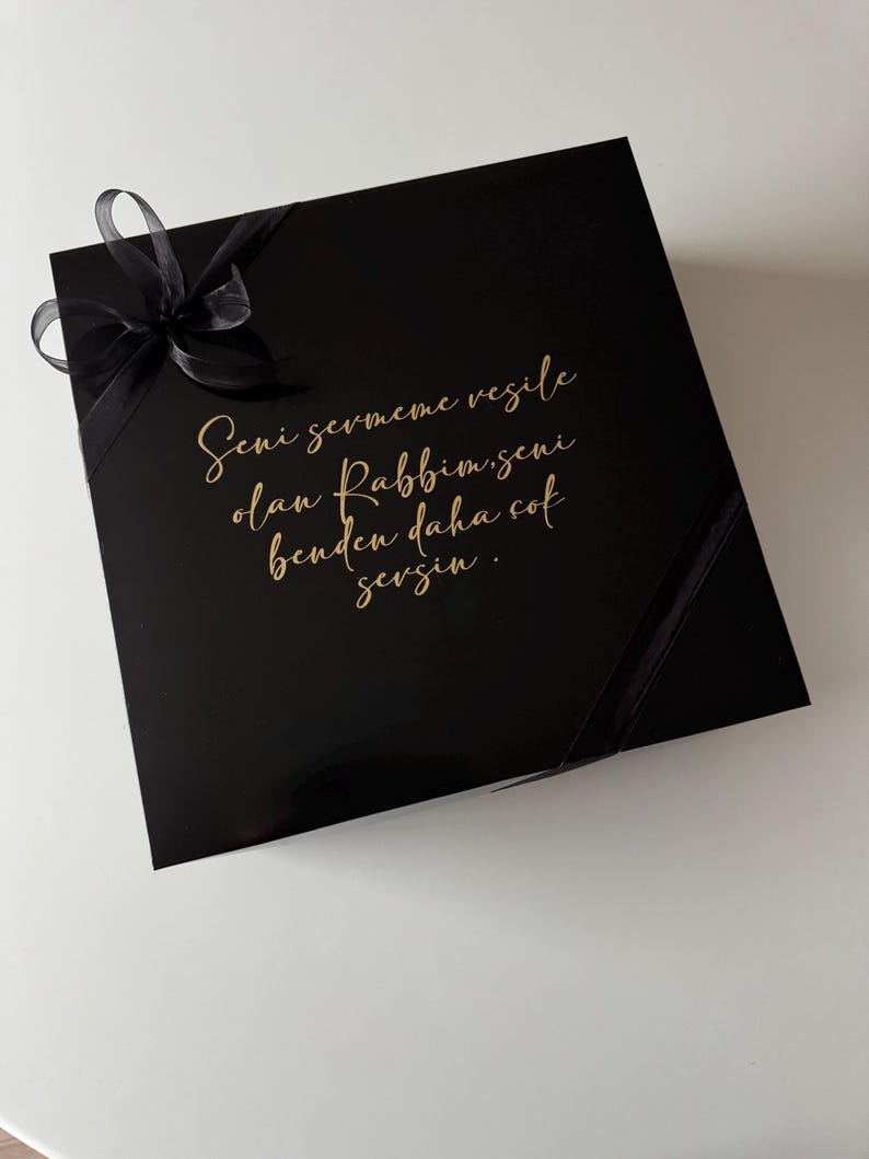 K&ouml;nnte beinhalten: Eine quadratische, schwarze Geschenkbox mit gl&auml;nzender Oberfl&auml;che, verziert mit einer schwarzen Schleife in der oberen linken Ecke. Der Deckel tr&auml;gt einen goldenen kursiven Schriftzug: Seni sevmeme vesile olan Rabbim, seni benden daha &ccedil;ok sevsin.