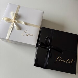 May include: A white square gift box with a cream ribbon and the name Esra in gold script, alongside a black square gift box with a black ribbon and the name Mevl&uuml;t in gold script. Both boxes feature a smooth finish and tied bows on top.