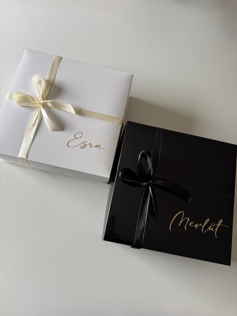 May include: A white square gift box with a cream ribbon and the name Esra in gold script, alongside a black square gift box with a black ribbon and the name Mevl&uuml;t in gold script. Both boxes feature a smooth finish and tied bows on top.