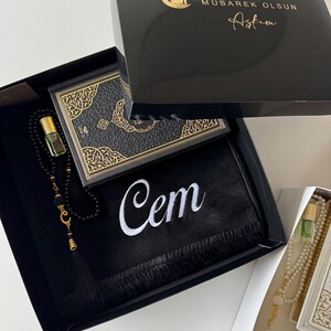 May include: A black gift box containing a black prayer mat with the name Cem embroidered in white, a black prayer bead strand with a gold crescent moon charm, a small glass perfume bottle, and a black book with gold ornate cover detailing.