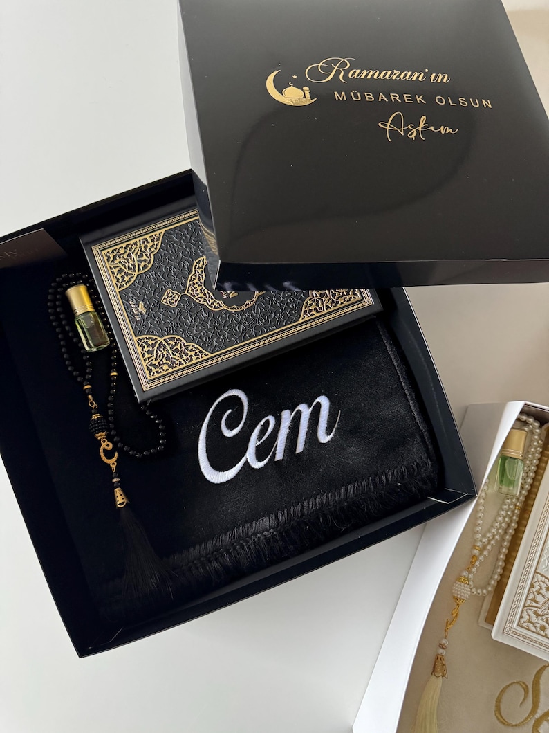 May include: A black gift box containing a black prayer mat with the name Cem embroidered in white, a black prayer bead strand with a gold crescent moon charm, a small glass perfume bottle, and a black book with gold ornate cover detailing.