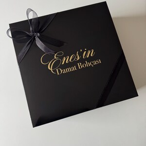 K&ouml;nnte beinhalten: Eine quadratische schwarze Geschenkbox mit einer schwarzen Schleife in der oberen linken Ecke. In der Mitte des Deckels befindet sich der goldene Schriftzug Enes'in Damat Boh&ccedil;ası in einer Kombination aus Schreib- und Serifenschrift.