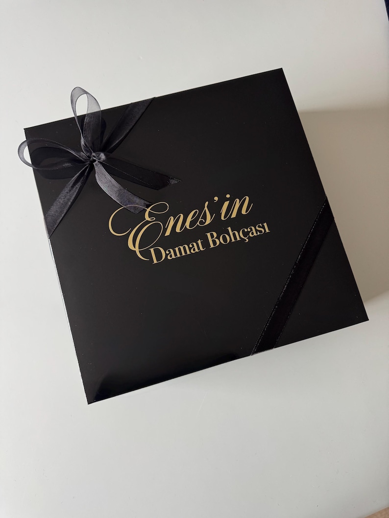 K&ouml;nnte beinhalten: Eine quadratische schwarze Geschenkbox mit einer schwarzen Schleife in der oberen linken Ecke. In der Mitte des Deckels befindet sich der goldene Schriftzug Enes'in Damat Boh&ccedil;ası in einer Kombination aus Schreib- und Serifenschrift.