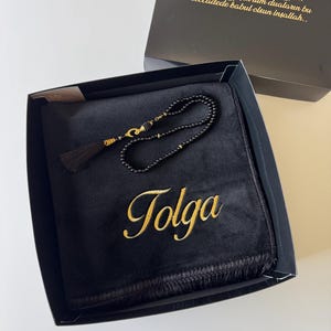 May include: A black prayer rug featuring the name Tolga embroidered in gold thread, accompanied by a set of black prayer beads with gold accents and a black tassel, all presented in a gift box with gold text printed on the inside lid.