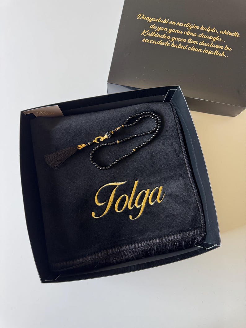 May include: A black prayer rug featuring the name Tolga embroidered in gold thread, accompanied by a set of black prayer beads with gold accents and a black tassel, all presented in a gift box with gold text printed on the inside lid.