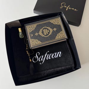 K&ouml;nnte beinhalten: Eine schwarze Geschenkbox mit einem schwarzen Gebetsteppich, auf dem der Name Safwan in Wei&szlig; gestickt ist, einem schwarz-goldenen Le Saint Coran Buch, einer schwarzen Gebetskette mit goldenen Akzenten und einem kleinen Glasfl&auml;schchen mit Parf&uuml;m&ouml;l.