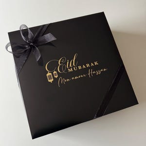 K&ouml;nnte beinhalten: Eine quadratische schwarze Geschenkbox mit einer goldenen Aufschrift Eid Mubarak Mon amour Hassan und zwei goldenen Laternen-Symbolen. Die Box ist mit einem transparenten schwarzen Band verziert, das oben links zu einer Schleife gebunden ist.