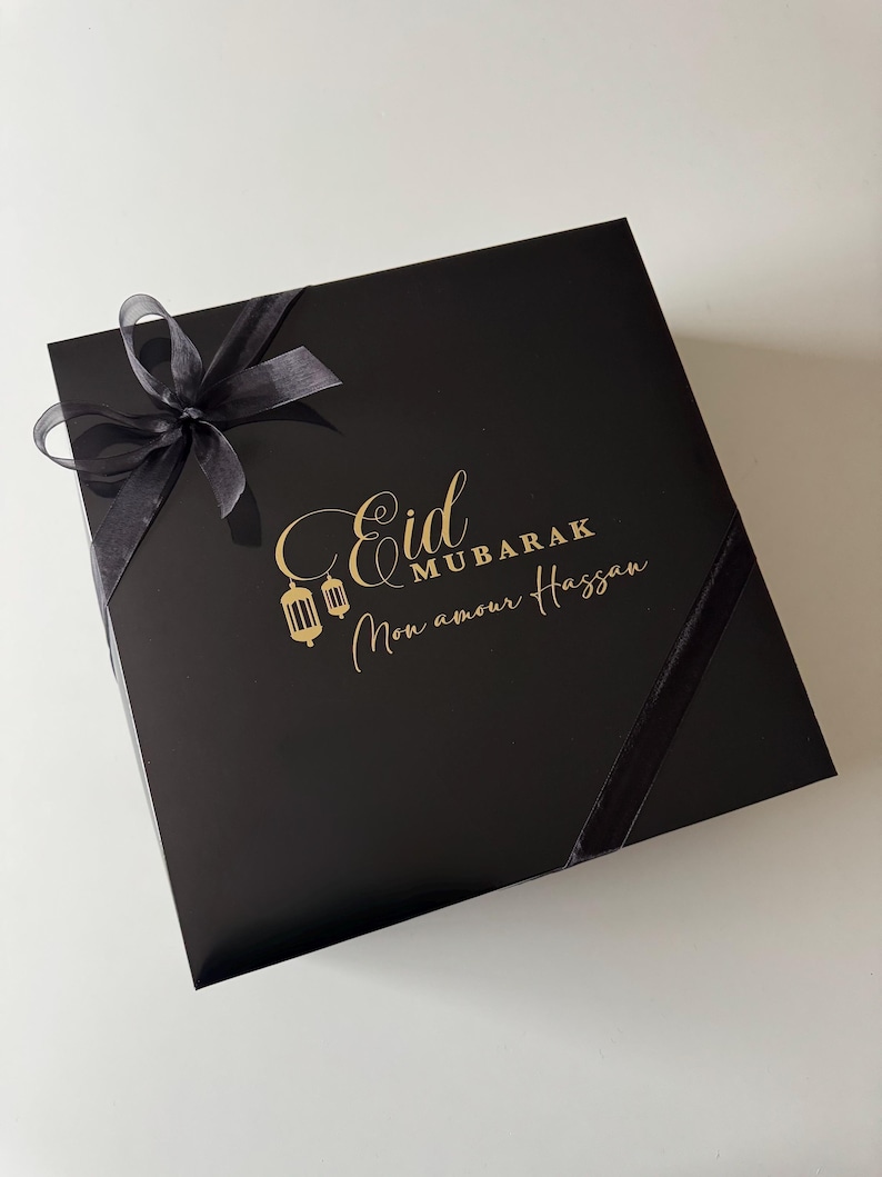 K&ouml;nnte beinhalten: Eine quadratische schwarze Geschenkbox mit einer goldenen Aufschrift Eid Mubarak Mon amour Hassan und zwei goldenen Laternen-Symbolen. Die Box ist mit einem transparenten schwarzen Band verziert, das oben links zu einer Schleife gebunden ist.