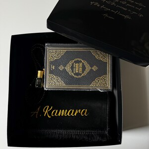 K&ouml;nnte beinhalten: Eine schwarze Geschenkbox mit einem kleinen Heiligen Koran mit goldgepr&auml;gtem Einband, einem schwarzen Gebetsteppich mit dem in Goldfaden gestickten Namen A. Kamara und einem kleinen Glasparf&uuml;mfl&auml;schchen. Der Deckel tr&auml;gt einen goldenen Text.