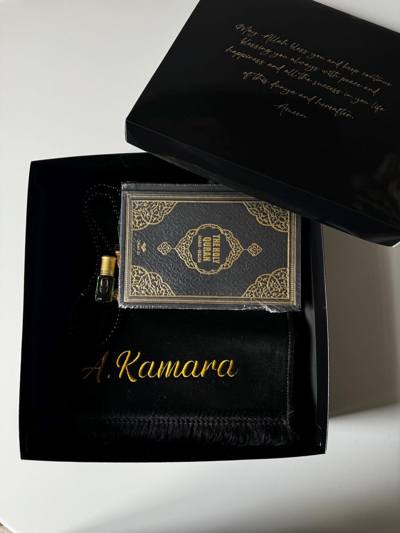 K&ouml;nnte beinhalten: Eine schwarze Geschenkbox mit einem kleinen Heiligen Koran mit goldgepr&auml;gtem Einband, einem schwarzen Gebetsteppich mit dem in Goldfaden gestickten Namen A. Kamara und einem kleinen Glasparf&uuml;mfl&auml;schchen. Der Deckel tr&auml;gt einen goldenen Text.