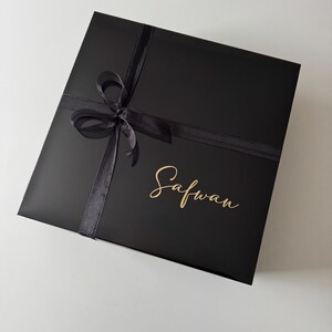 K&ouml;nnte beinhalten: Eine quadratische schwarze Geschenkbox mit einer schwarzen Schleife auf der Oberseite. Der Name Safwan ist in goldener Schreibschrift auf dem Deckel der Box aufgedruckt. Die Box hat eine glatte, matte Oberfl&auml;che und ein schlichtes Design.
