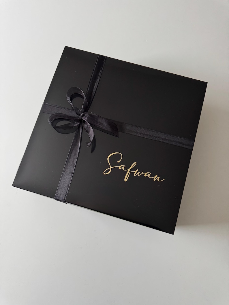 K&ouml;nnte beinhalten: Eine quadratische schwarze Geschenkbox mit einer schwarzen Schleife auf der Oberseite. Der Name Safwan ist in goldener Schreibschrift auf dem Deckel der Box aufgedruckt. Die Box hat eine glatte, matte Oberfl&auml;che und ein schlichtes Design.