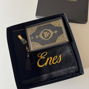 K&ouml;nnte beinhalten: Eine schwarze Geschenkbox mit einem schwarzen Gebetsteppich, der mit dem Namen Enes in Goldfaden bestickt ist, einem kleinen schwarzen Buch mit dem Titel Le Saint Coran mit goldenen Zierr&auml;ndern und einer schwarzen Gebetskette mit goldener Quaste.
