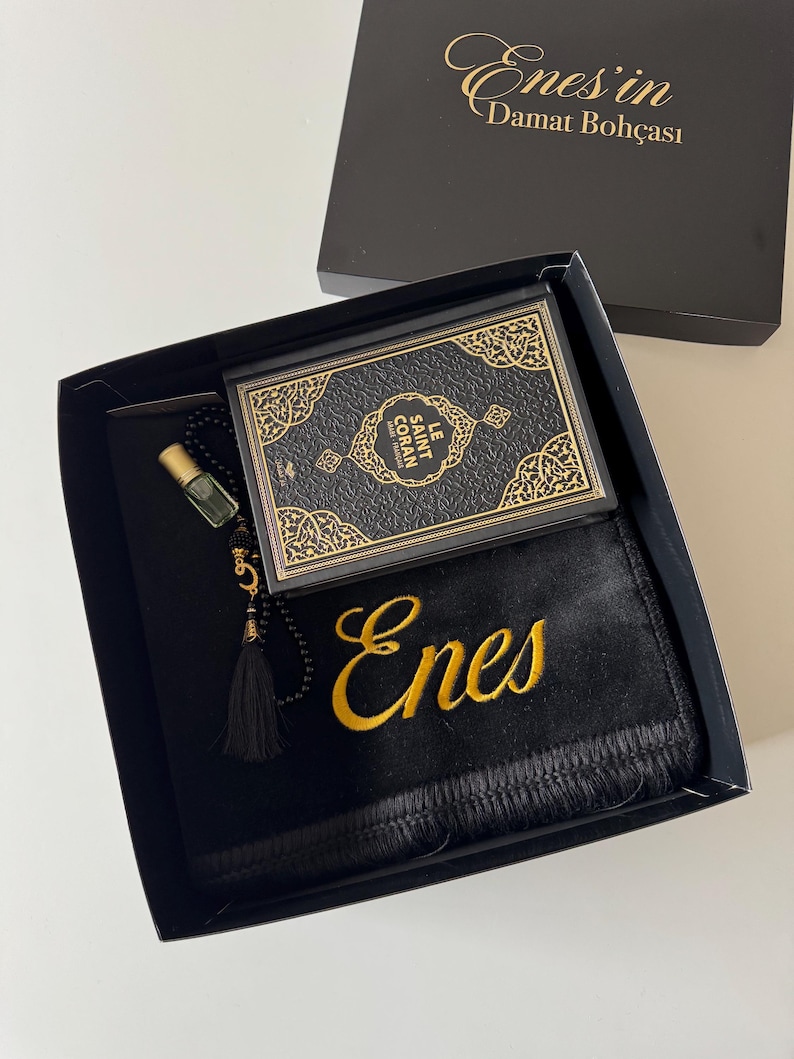 K&ouml;nnte beinhalten: Eine schwarze Geschenkbox mit einem schwarzen Gebetsteppich, der mit dem Namen Enes in Goldfaden bestickt ist, einem kleinen schwarzen Buch mit dem Titel Le Saint Coran mit goldenen Zierr&auml;ndern und einer schwarzen Gebetskette mit goldener Quaste.