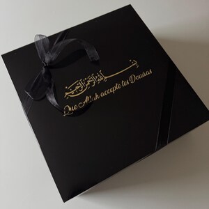 K&ouml;nnte beinhalten: Eine quadratische schwarze Geschenkbox mit einer schwarzen Schleife in der oberen linken Ecke. Der Deckel zeigt goldene arabische Kalligrafie &uuml;ber dem goldenen Text "Que Allah accepte tes Dou&acirc;as" in einer kursiven Schriftart.