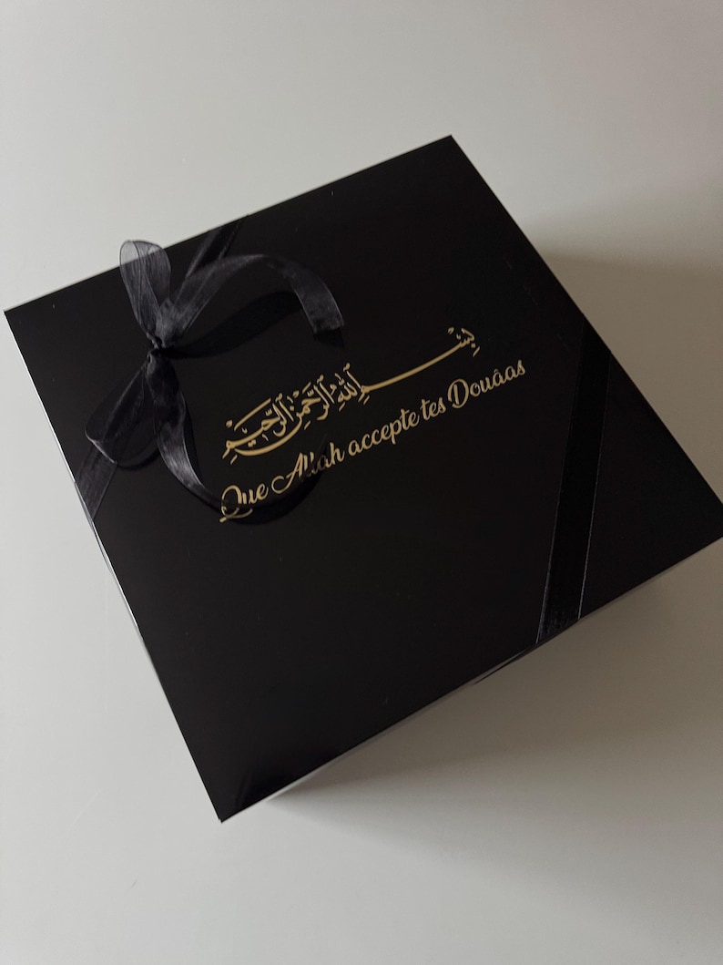 K&ouml;nnte beinhalten: Eine quadratische schwarze Geschenkbox mit einer schwarzen Schleife in der oberen linken Ecke. Der Deckel zeigt goldene arabische Kalligrafie &uuml;ber dem goldenen Text "Que Allah accepte tes Dou&acirc;as" in einer kursiven Schriftart.