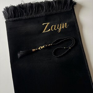 K&ouml;nnte beinhalten: Ein schwarzer Gebetsteppich mit Fransen am oberen Rand und dem in Goldfaden gestickten Namen Zayn. Auf dem Teppich liegt eine Kette aus schwarzen Gebetsperlen mit goldfarbenen Akzenten und einer schwarzen Quaste, die kreisf&ouml;rmig angeordnet ist.