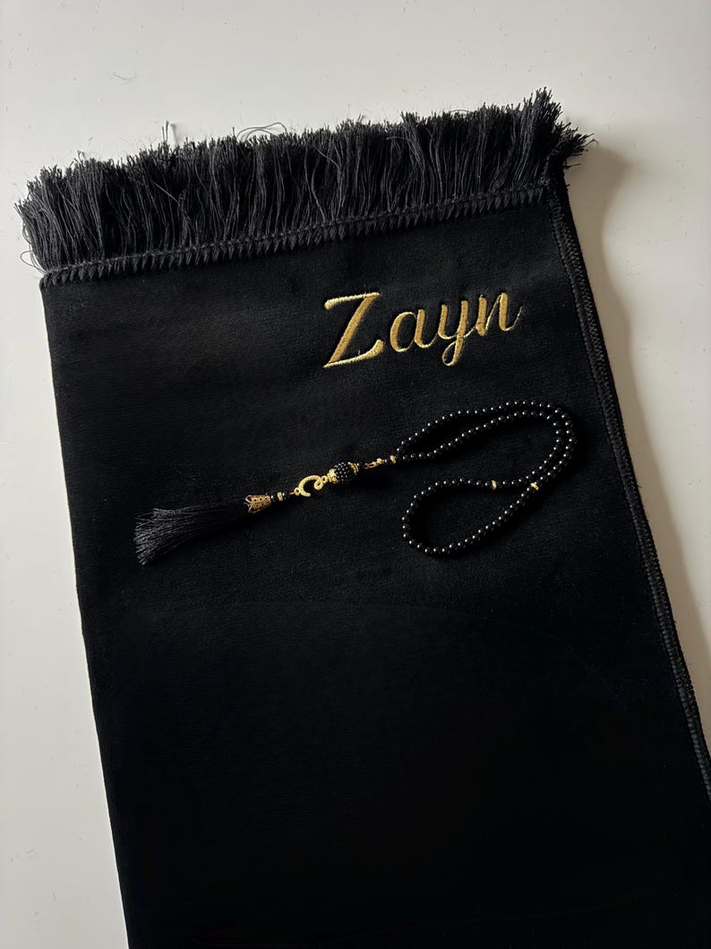 K&ouml;nnte beinhalten: Ein schwarzer Gebetsteppich mit Fransen am oberen Rand und dem in Goldfaden gestickten Namen Zayn. Auf dem Teppich liegt eine Kette aus schwarzen Gebetsperlen mit goldfarbenen Akzenten und einer schwarzen Quaste, die kreisf&ouml;rmig angeordnet ist.